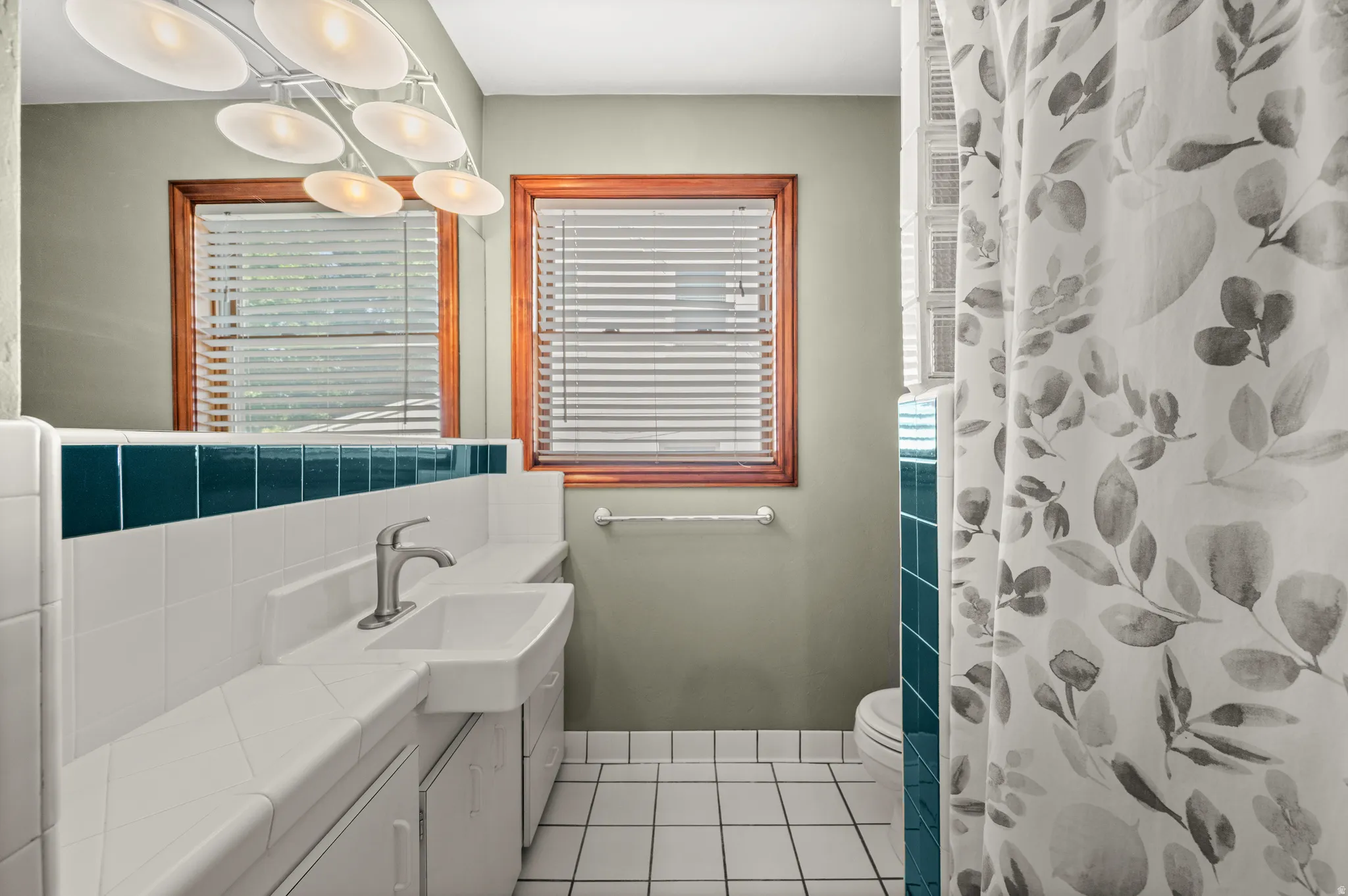 Bathroom featuring a white tile floor, white tiled vanity with a single basin sink, a wall-mounted mirror, and two windows with wood trim
