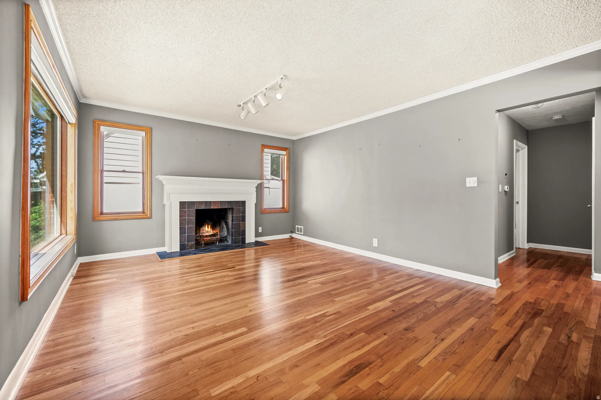 Spacious room featuring wood-finish flooring, a white mantel fireplace with a dark tile surround, multiple windows with wood trim, and track lighting