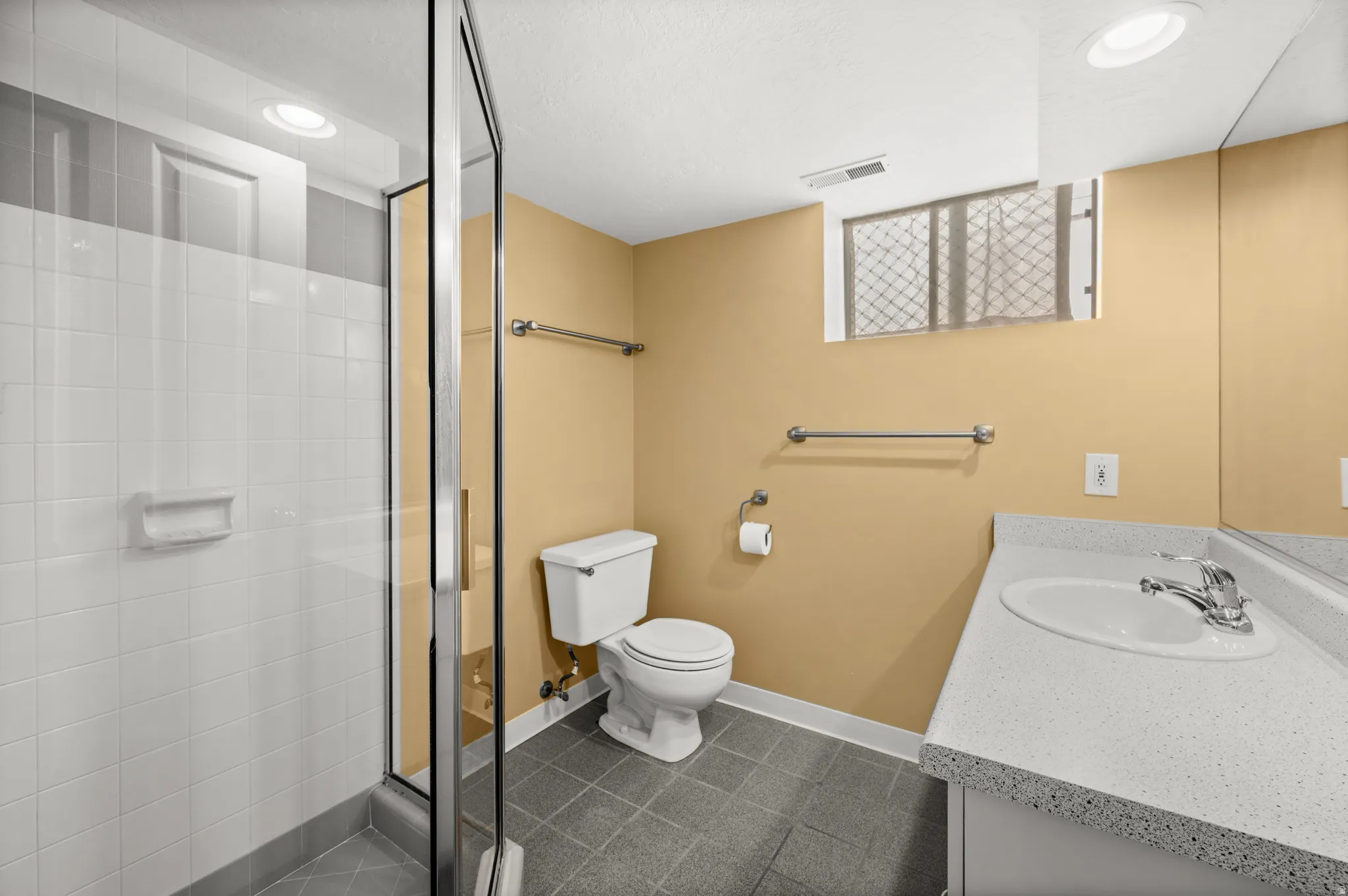 Bathroom featuring a glass-enclosed shower with white tiled walls, a single vanity with a white countertop and integrated sink, a toilet, and gray tiled flooring