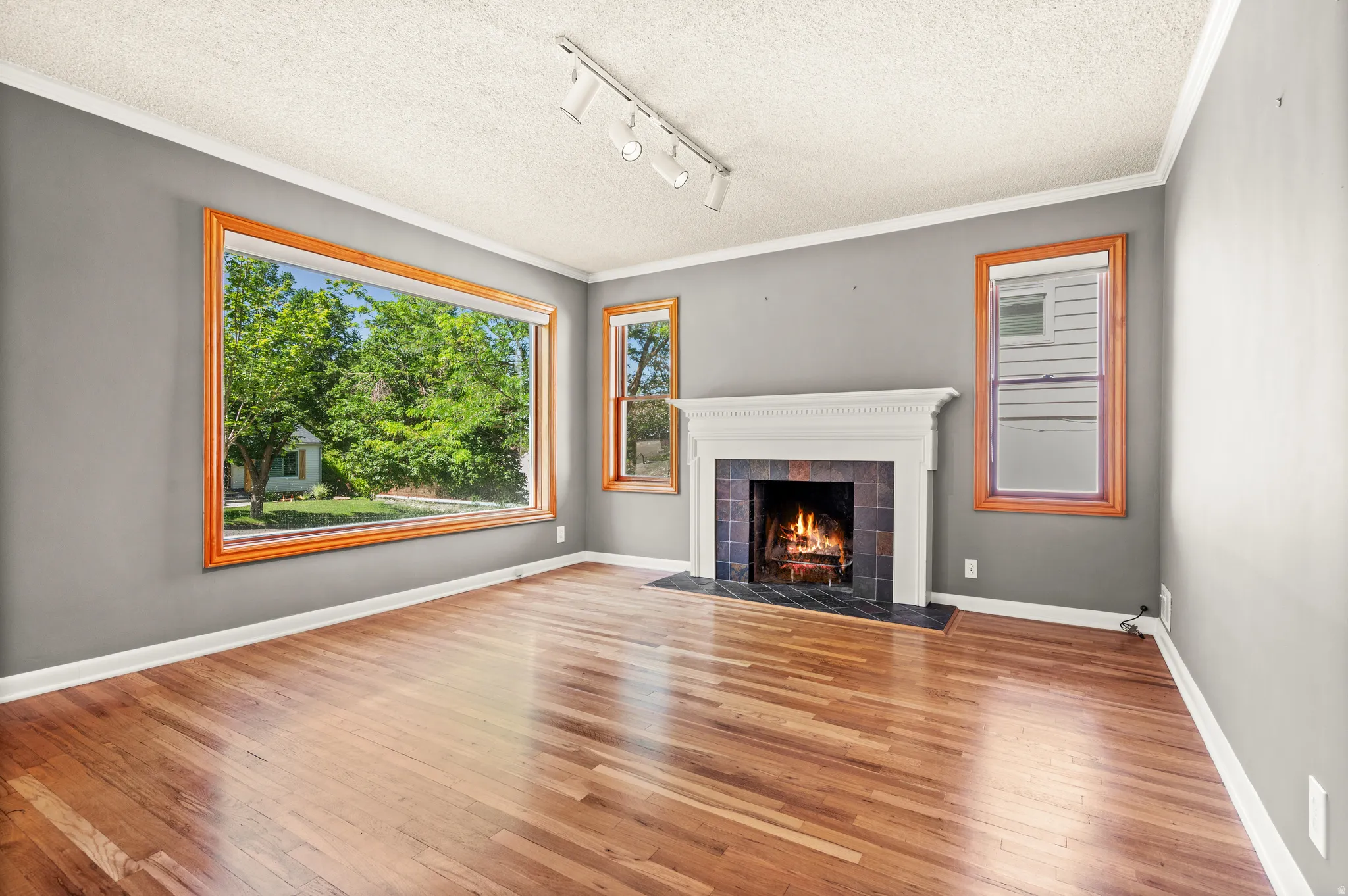 Spacious living area featuring wood-finish flooring, a traditional fireplace with a white mantel and dark tile surround, and large windows with natural wood trim