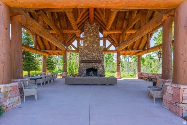 Expansive covered patio featuring a stone fireplace, exposed timber beams, a vaulted wood ceiling, concrete flooring, and stone pillar bases