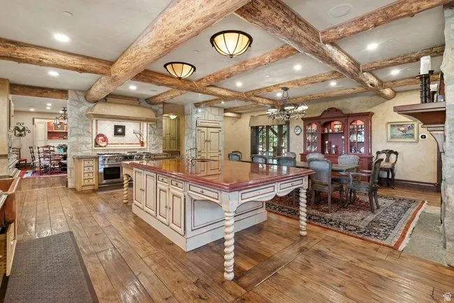 Expansive kitchen and dining area featuring wood-finish flooring, exposed wood beam ceiling, central island with storage and integrated sink, stainless steel range, and stone accents