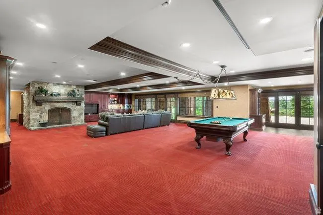 Spacious recreation room featuring a stone fireplace with hearth, red patterned carpeting, recessed lighting, and dark wood ceiling beams