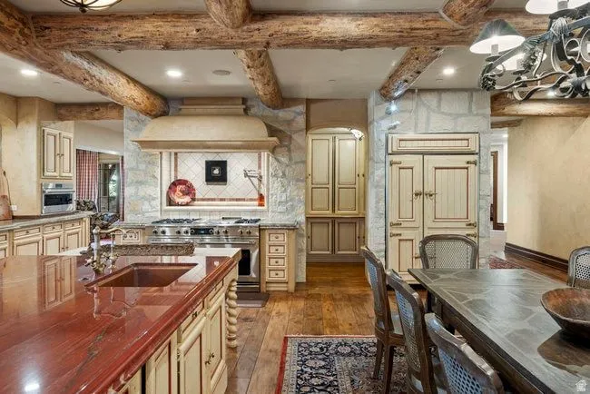 Gourmet kitchen featuring a large wood-finish island with an integrated sink, custom cabinetry with ornate hardware, professional-grade stainless steel range with custom hood, stone accent walls, and exposed wood beams