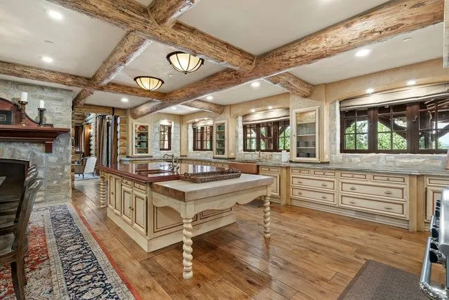 Expansive kitchen featuring exposed wood beam ceilings, wood-finish flooring, a substantial center island with butcher block and stone countertops, custom cabinetry with glass-front display units, and stone wall accents