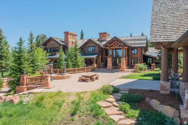 Expansive log home with stone chimneys, featuring a flagstone patio, built-in fire pit, and mature evergreen landscaping
