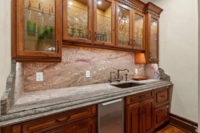 Built-in wet bar featuring custom wood cabinetry with glass-front uppers, under-cabinet lighting, a stone countertop, and a matching stone backsplash