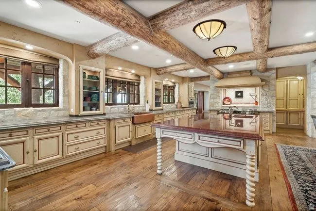 Rustic kitchen featuring exposed wood beams, light-toned custom cabinetry, wood-finish flooring, a central island with a polished countertop, and a built-in copper farmhouse sink