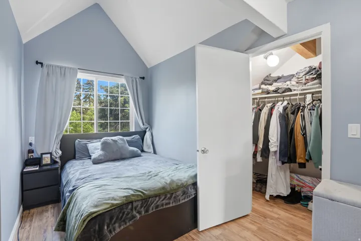 Bedroom featuring a vaulted ceiling with exposed beams, a large grid-pattern window, wood-finish flooring, and a built-in closet with shelving and hanging rods