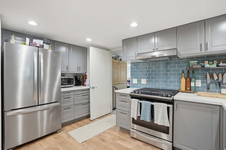 Modern kitchen featuring light gray cabinetry, stainless steel appliances, a blue subway tile backsplash, and light wood-finish flooring