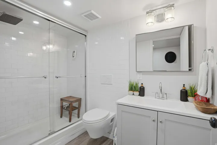 Bathroom featuring a glass-enclosed shower with subway tile surround, a wall-mounted toilet, a vanity with a white countertop and integrated sink, and wood-finish flooring