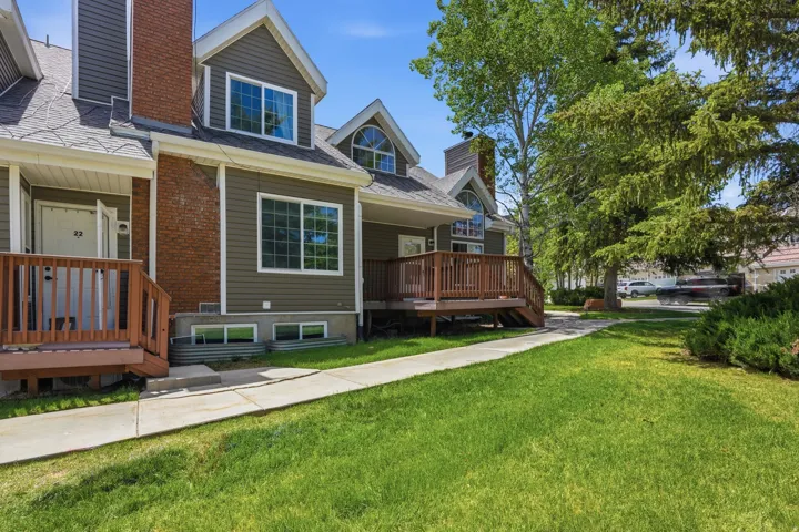 Exterior featuring dark green siding, brick chimney accents, gabled dormers, a wood deck with railings, and established perimeter trees