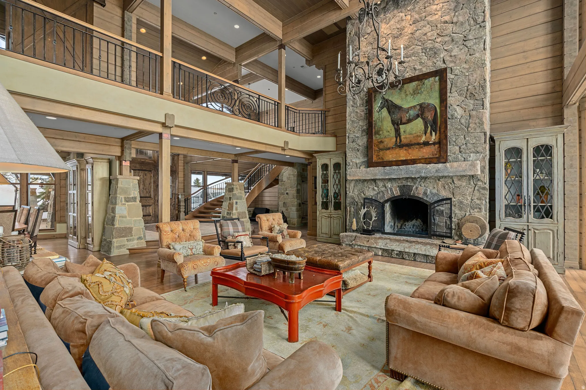 Great room featuring a floor-to-ceiling stone fireplace, exposed wood beams, a second-story balcony with wrought iron railing, wood-paneled walls, and a grand staircase