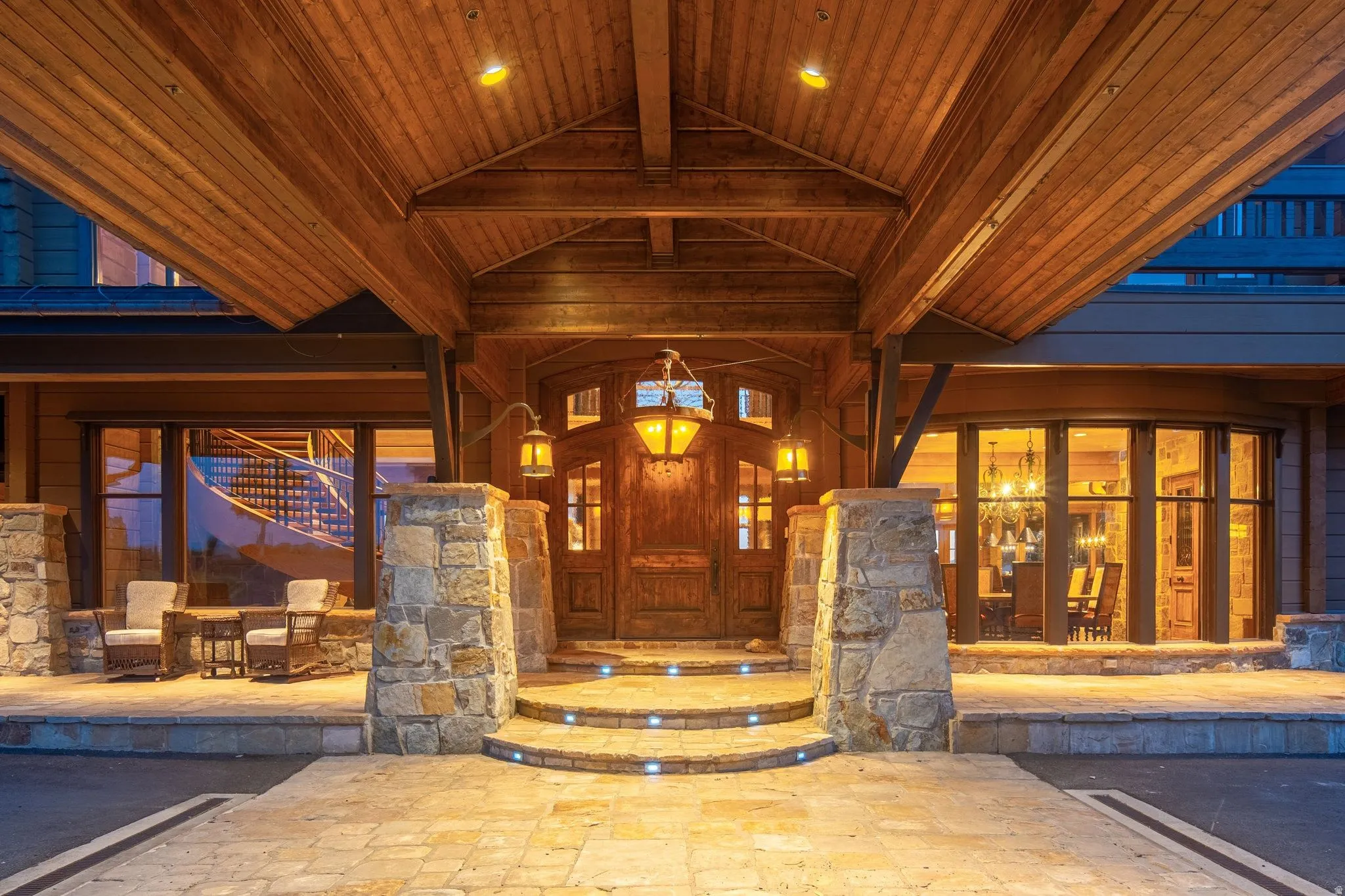 Stone and timber entry featuring a covered porte-cochère, decorative double doors, integrated step lighting, a stone-faced foundation, and an outdoor patio