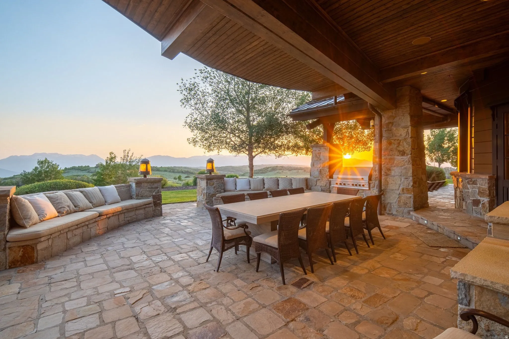 Expansive stone patio featuring built-in seating, a stone outdoor kitchen with a grill, and a wood-paneled ceiling