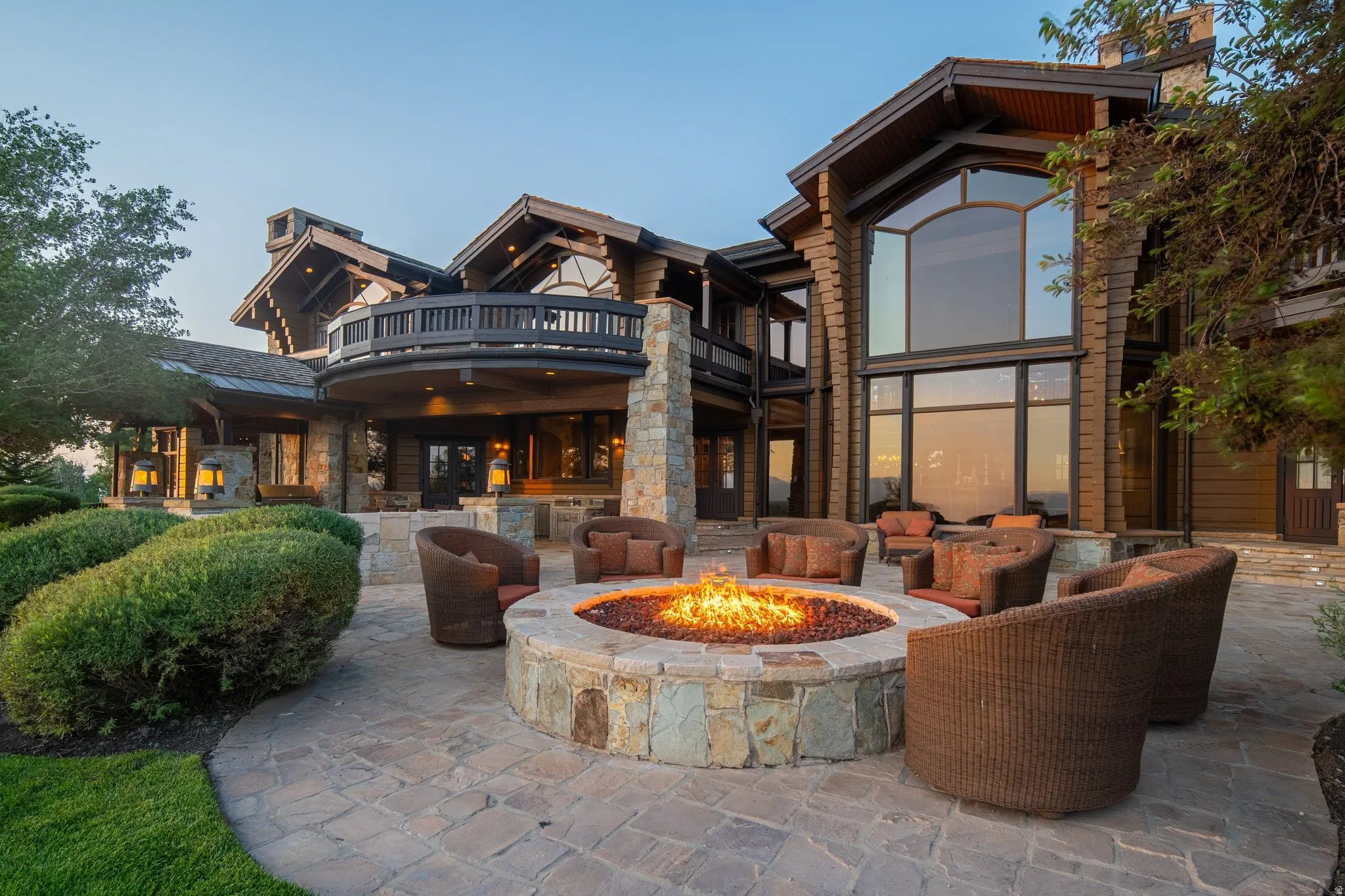 Expansive stone patio with a built-in stone fire pit, a curved upper-level balcony, large picture windows, and a log cabin style exterior