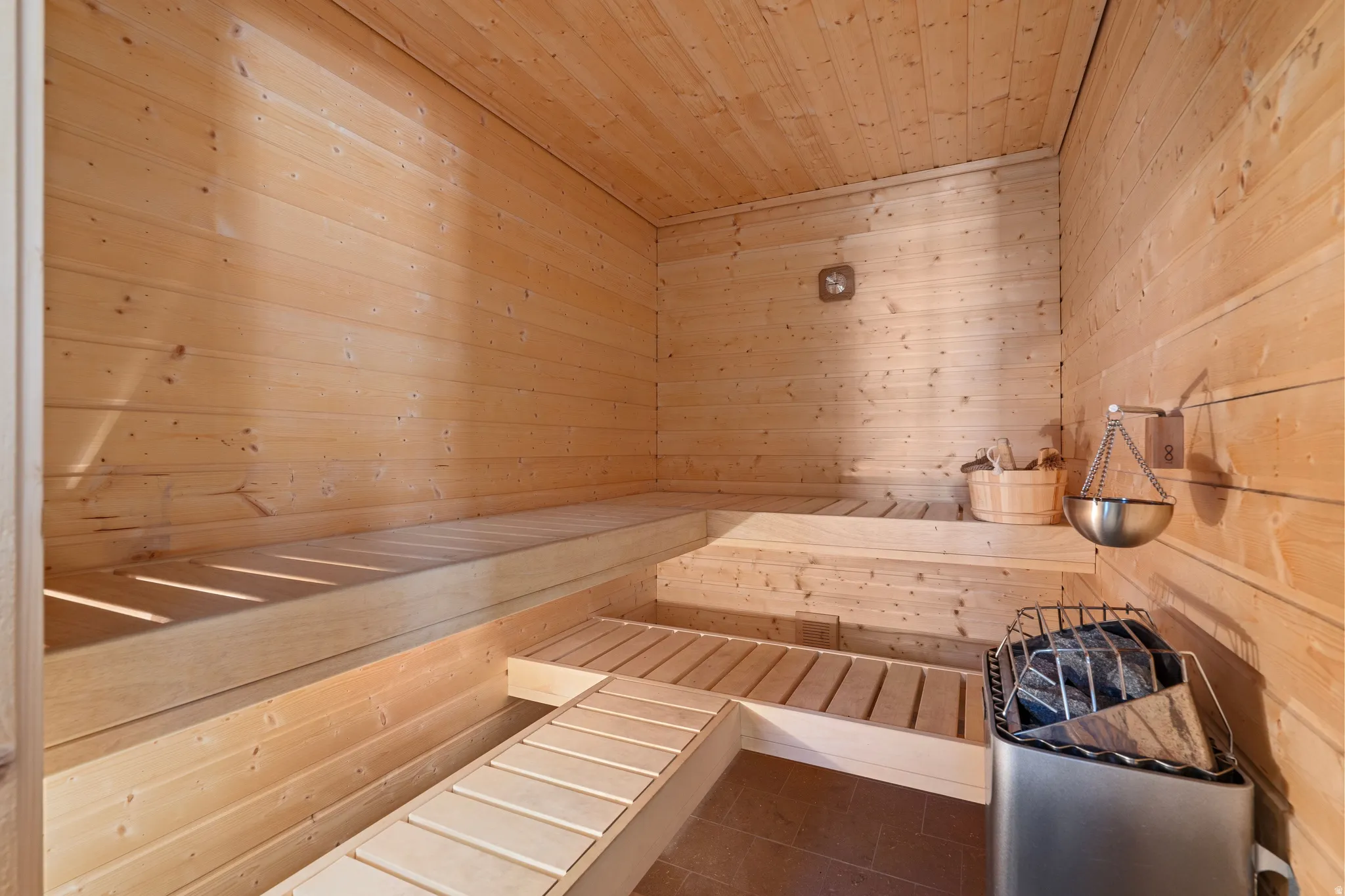 Sauna featuring natural wood paneling, multi-level seating, slatted flooring, a stainless steel heater, and a wall-mounted water basin
