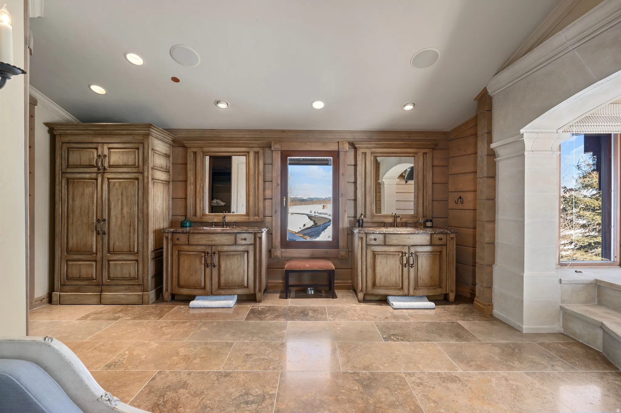 Spacious room featuring extensive stone tile flooring, custom wood cabinetry, dual vanity areas with integrated sinks, and ample natural light from multiple windows