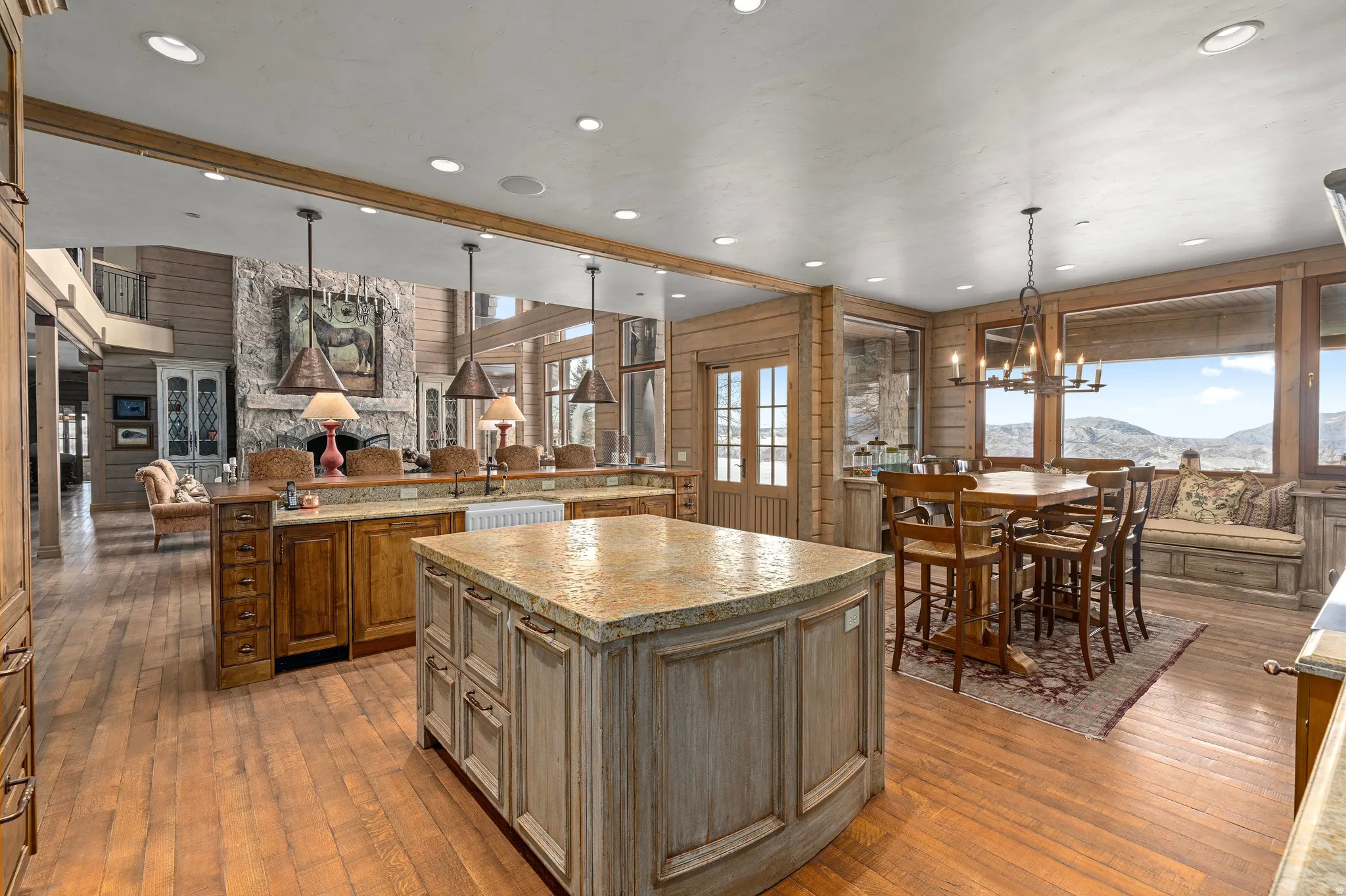 Expansive kitchen and dining area featuring wood-finish flooring, two large kitchen islands with stone countertops, a farmhouse sink, and panoramic windows showcasing mountain scenery
