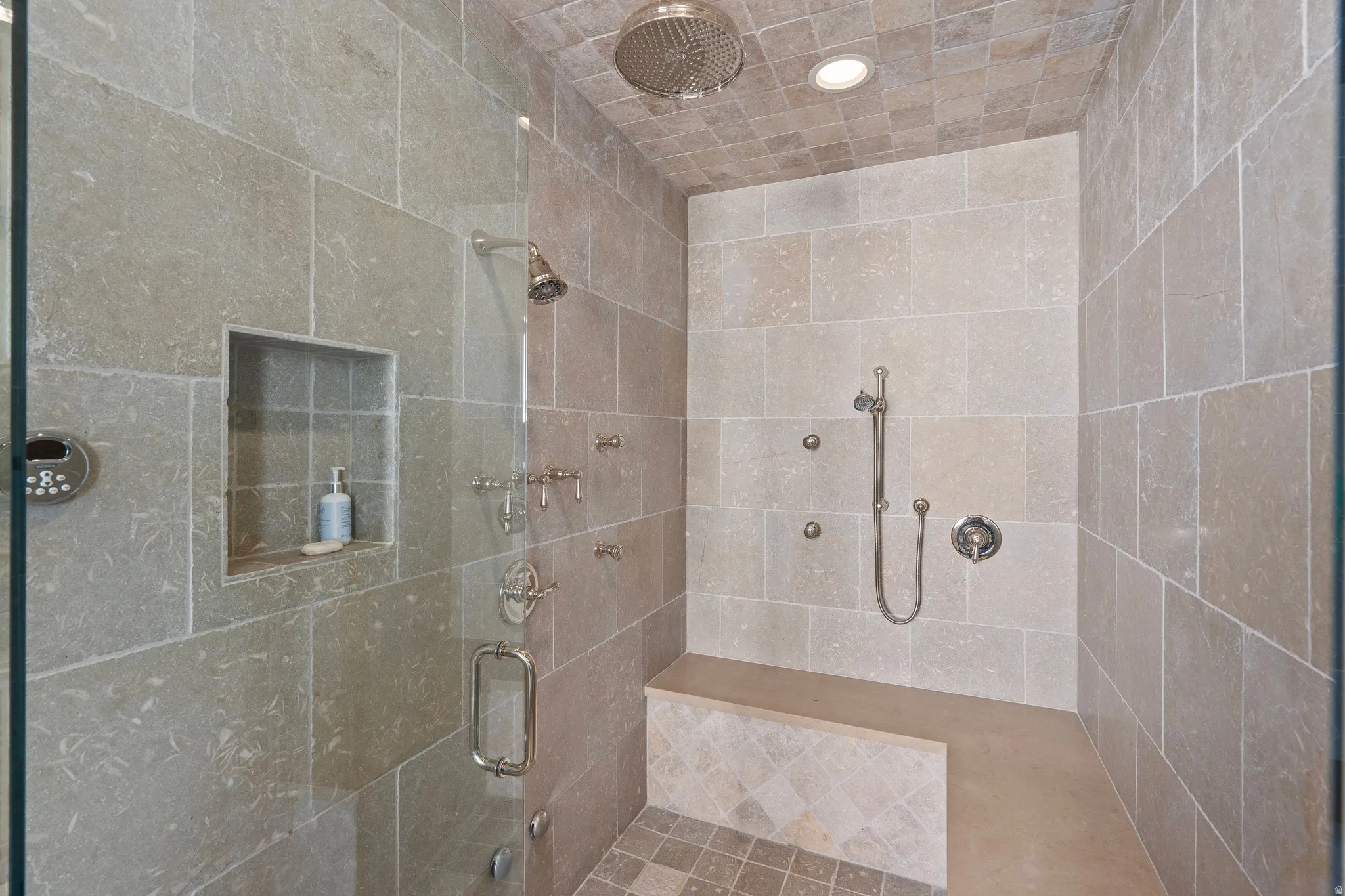Shower enclosure featuring full-height stone-look tile, a built-in bench, a recessed niche, multiple showerheads, and a glass door with a polished chrome handle