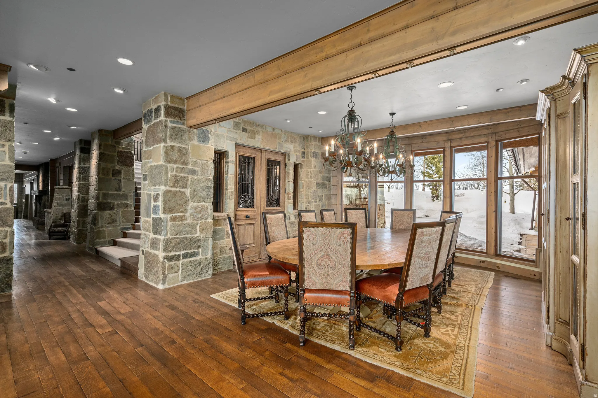 Spacious dining area featuring wood-finish flooring, stone accent walls, exposed ceiling beams, and a prominent chandelier