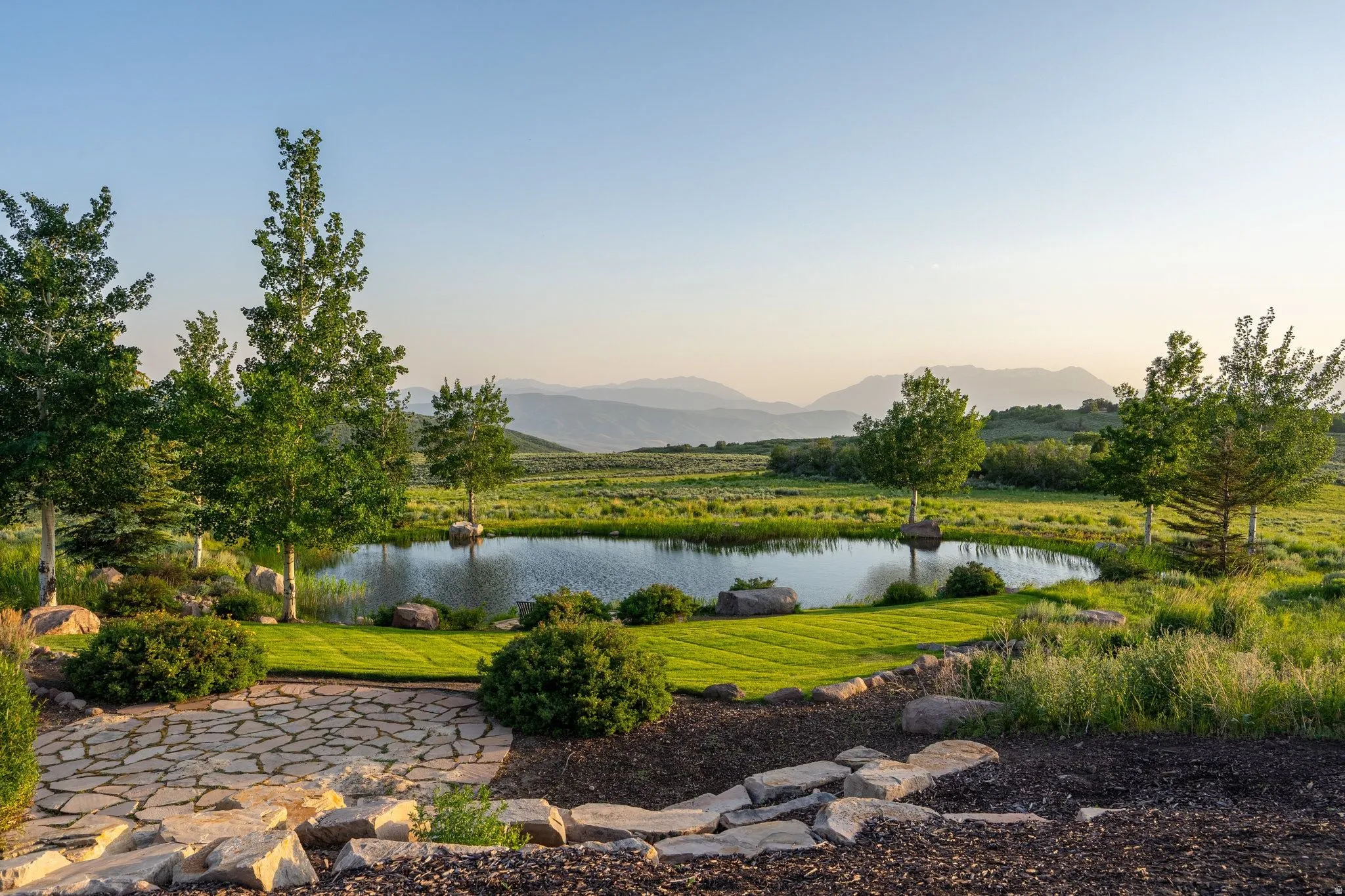 Manicured lawn surrounding a private pond with established trees, a flagstone patio, and mountainous terrain in the distance