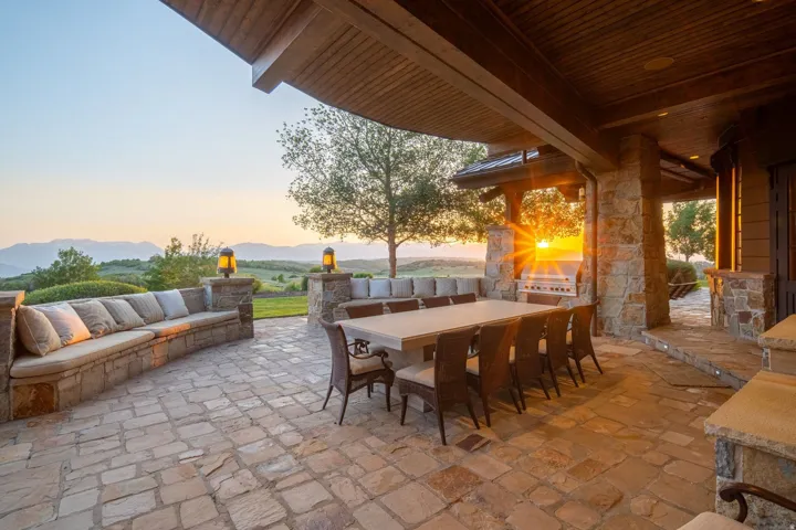 Expansive stone patio featuring built-in seating, a stone outdoor kitchen with a grill, and a wood-paneled ceiling