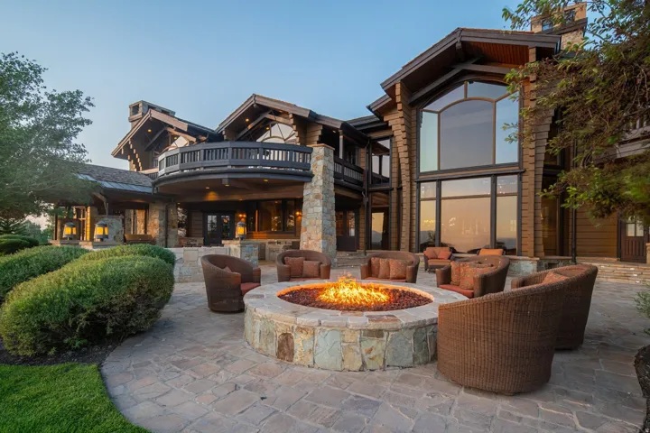 Expansive stone patio with a built-in stone fire pit, a curved upper-level balcony, large picture windows, and a log cabin style exterior