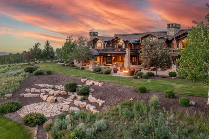 Expansive multi-level property featuring a stone and timber exterior, multiple balconies, a spacious stone patio, an outdoor fire pit area, and landscaped grounds with mature trees and stone hardscaping