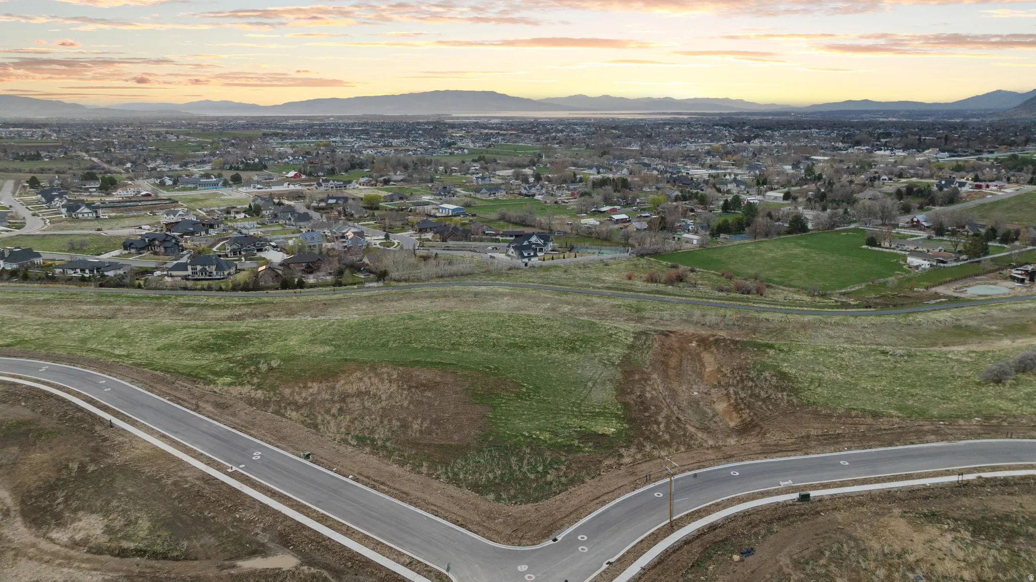 Expansive undeveloped land featuring a paved road with concrete sidewalks, established infrastructure, and distant residential areas