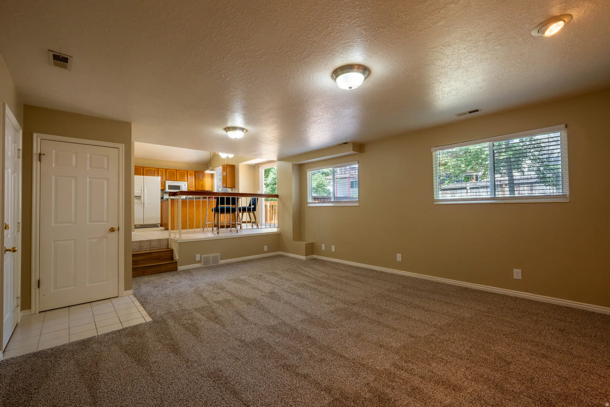 Spacious carpeted living area with two windows, recessed lighting, and a raised kitchen featuring wood cabinetry, white appliances, and a breakfast bar