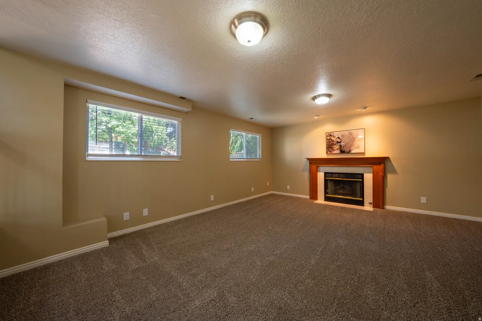Spacious room featuring neutral carpeting, a fireplace with a wood mantle and tile surround, two windows with blinds, and recessed lighting