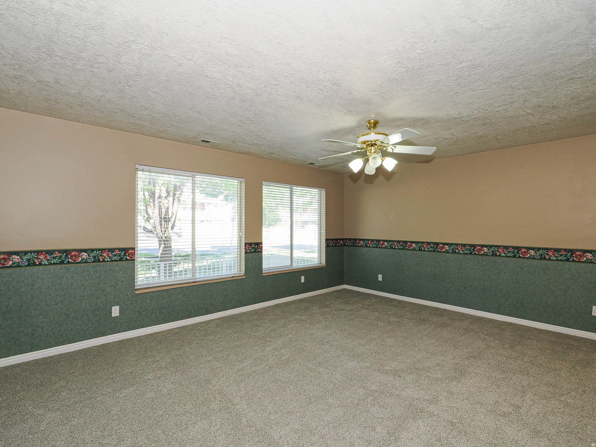 Carpeted interior space featuring two large windows with blinds, a ceiling fan with integrated lighting, and walls with a decorative border and wainscoting