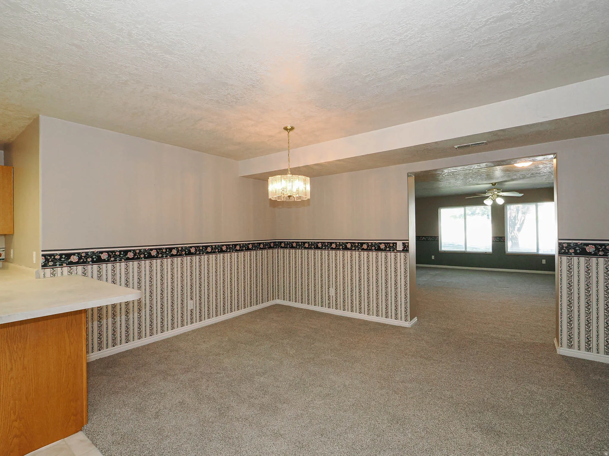 Carpeted living area featuring a rectangular chandelier, a partial breakfast bar with a light-toned countertop, and an open doorway to an additional room with multiple windows and a ceiling fan