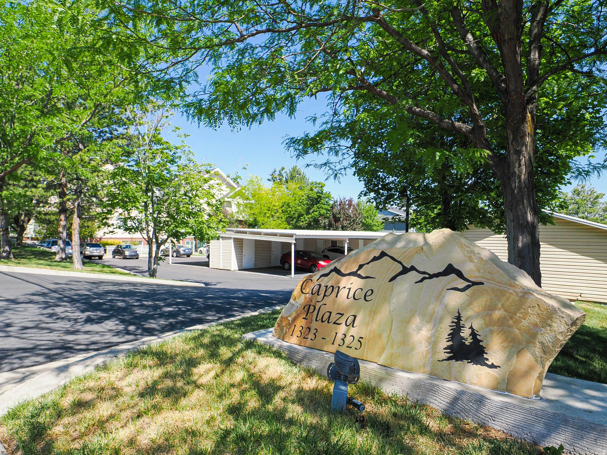 Property entrance featuring a stone monument sign, mature trees, and covered parking structures