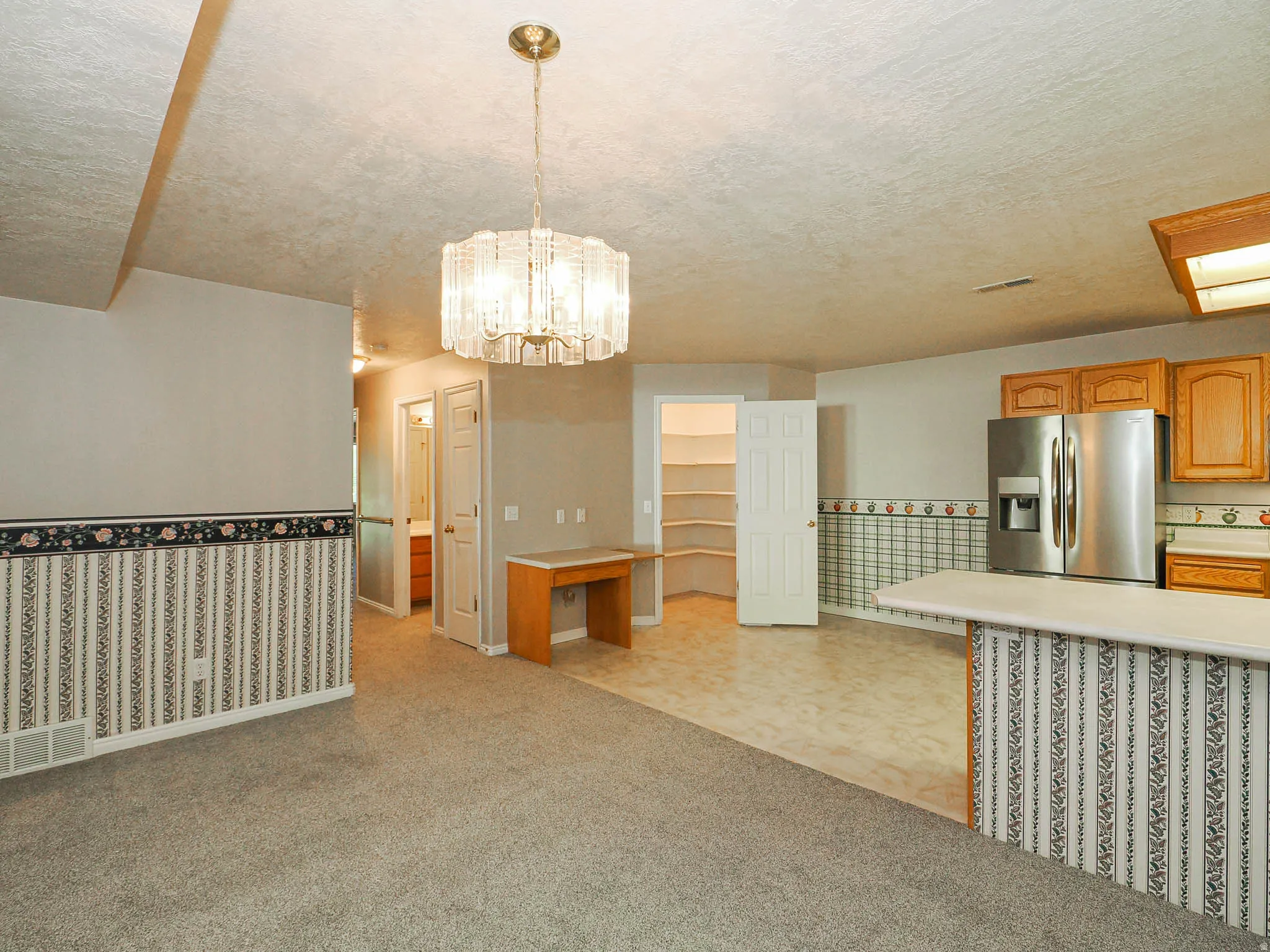 Open-concept living area featuring a central chandelier, a built-in desk, and a kitchen area with wood-finish cabinetry, stainless steel refrigerator, and a breakfast bar with a solid surface countertop