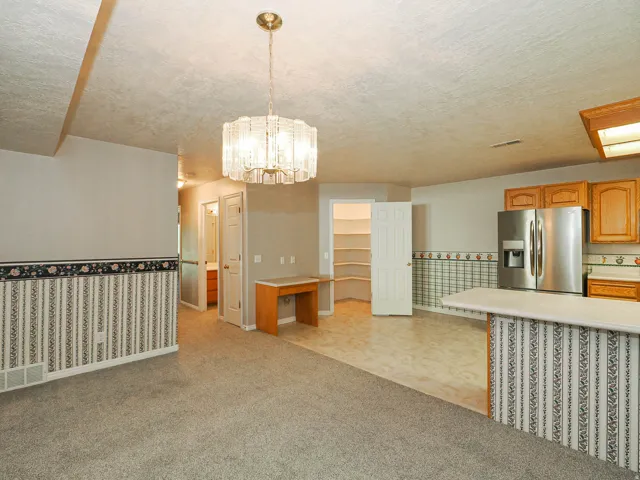 Open-concept living area featuring a central chandelier, a built-in desk, and a kitchen area with wood-finish cabinetry, stainless steel refrigerator, and a breakfast bar with a solid surface countertop