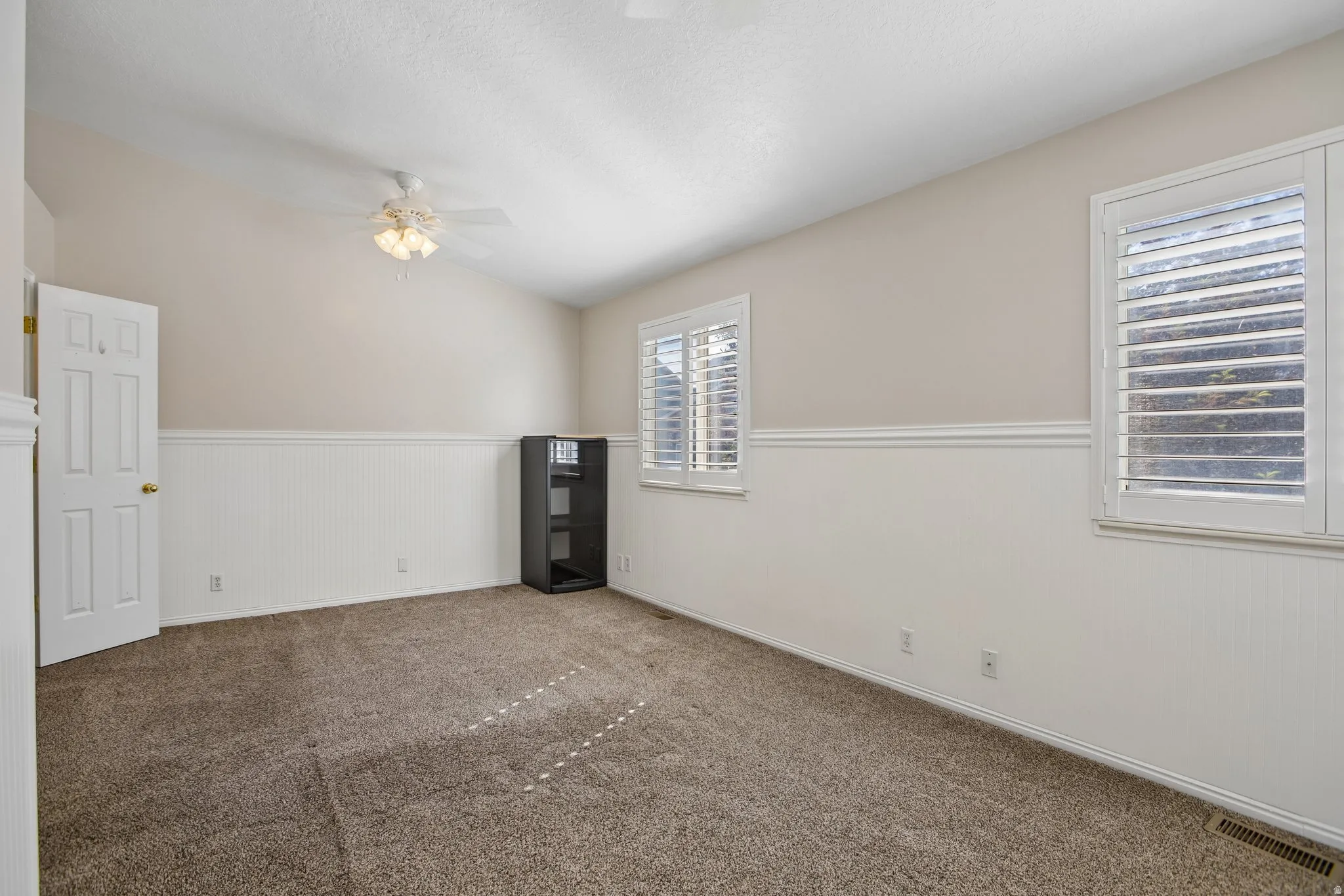 Carpeted room featuring a ceiling fan, two windows with plantation shutters, white wainscoting, and neutral-toned walls