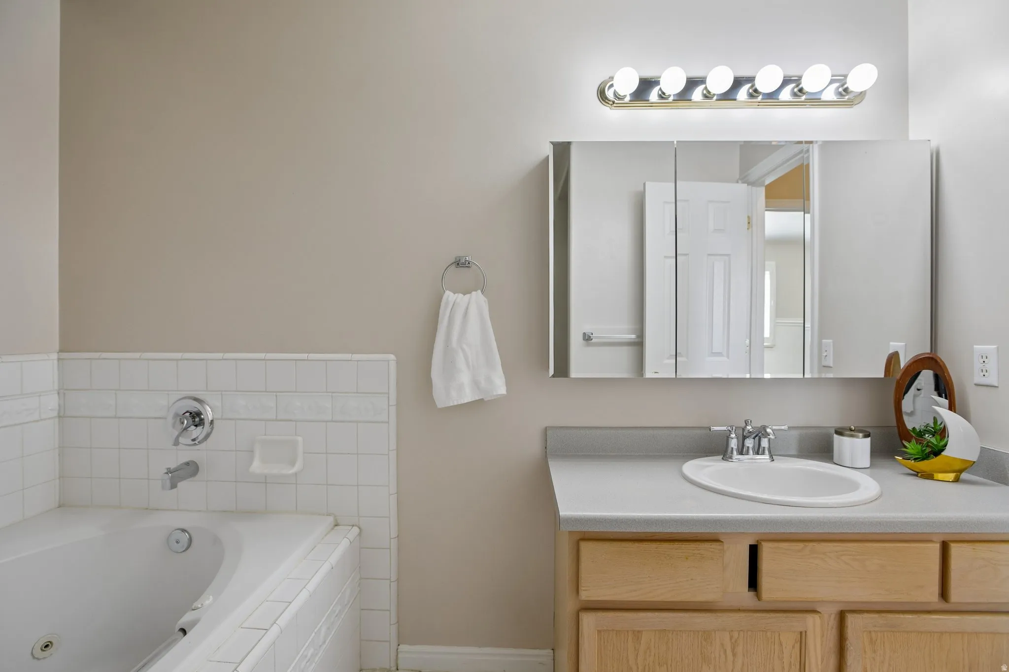 Bathroom featuring a white tiled soaking tub, a vanity with a laminate countertop, an integrated sink, natural wood-finish cabinetry, and a mirrored medicine cabinet with overhead lighting