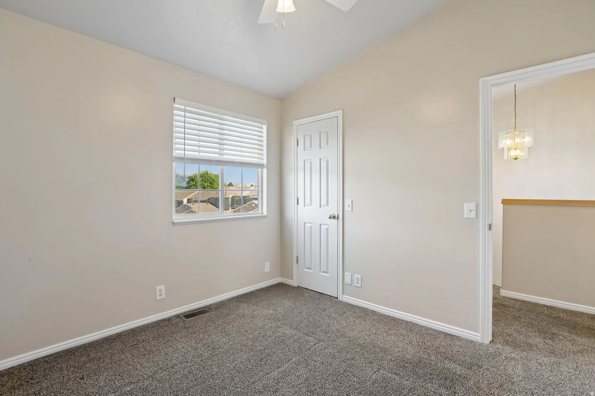 Vaulted ceiling room featuring neutral-toned walls, gray textured carpeting, a single window with horizontal blinds, and a white paneled door