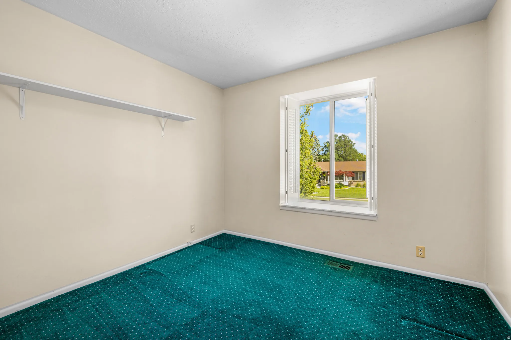 Room featuring a white-framed window with louvered shutters, light beige walls, and teal patterned carpeting