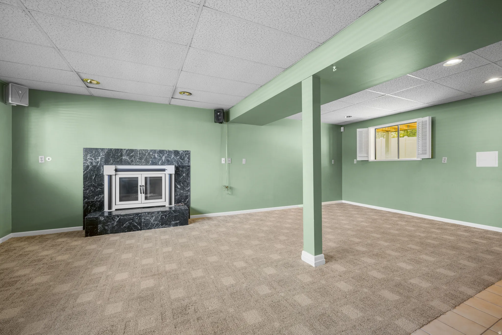Spacious room featuring a dark stone fireplace with a glass enclosure, patterned carpet, light green walls, a drop ceiling with recessed lighting, and a window with white louvered shutters