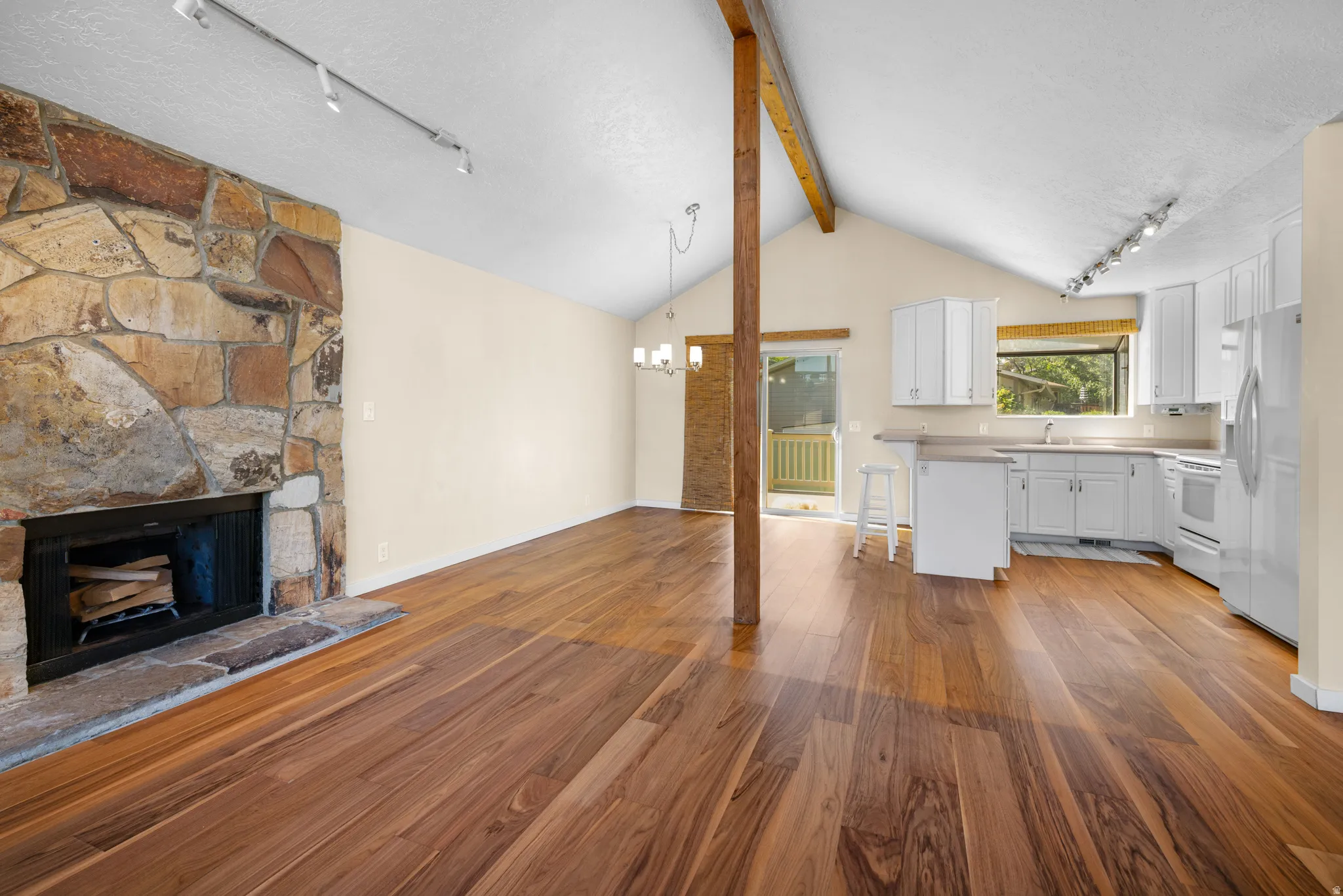 Vaulted ceiling interior featuring wood-finish flooring, a stone-faced fireplace, exposed beam, white cabinetry, and a sliding glass door leading to an exterior balcony