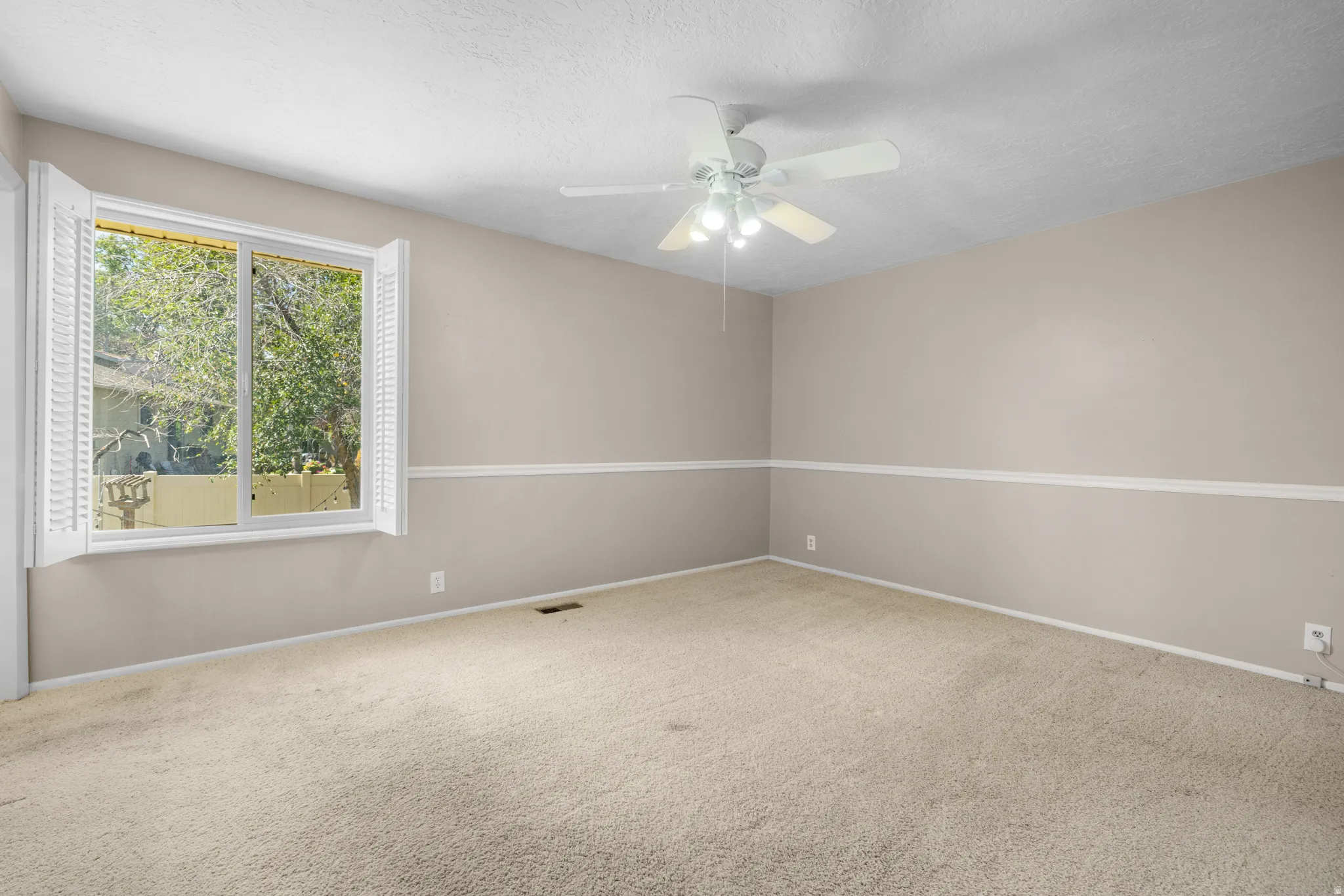 Spacious room featuring neutral carpeting, light beige walls, white chair rail molding, a large window with white plantation shutters, and a ceiling fan with integrated lighting