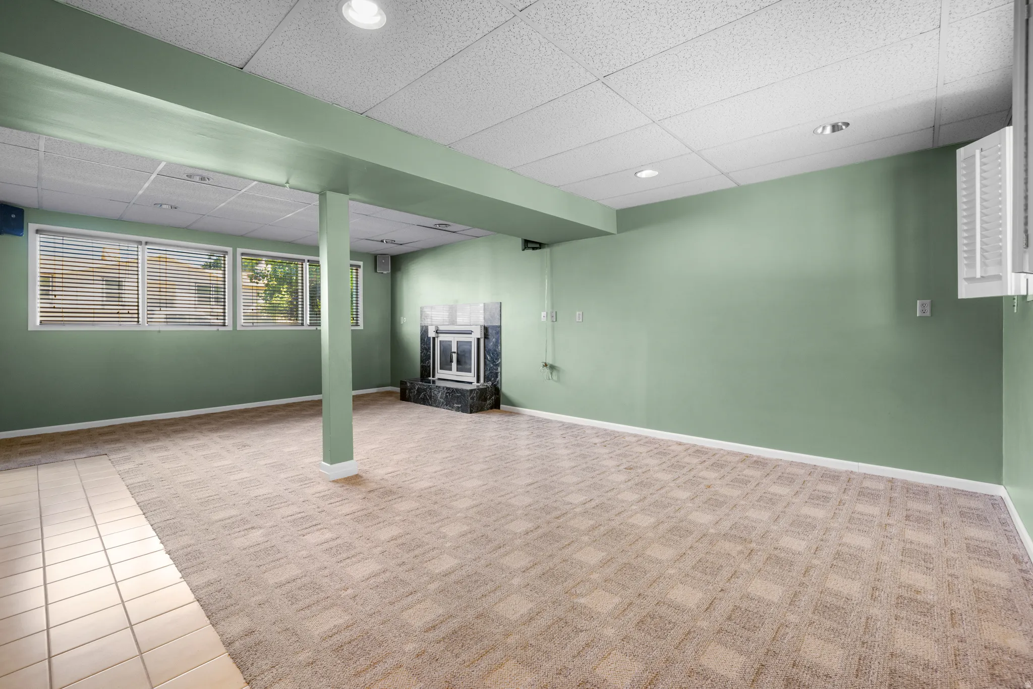 Spacious room featuring a corner fireplace with a dark stone surround, multiple windows with blinds, recessed lighting, and a combination of patterned carpet and tile flooring