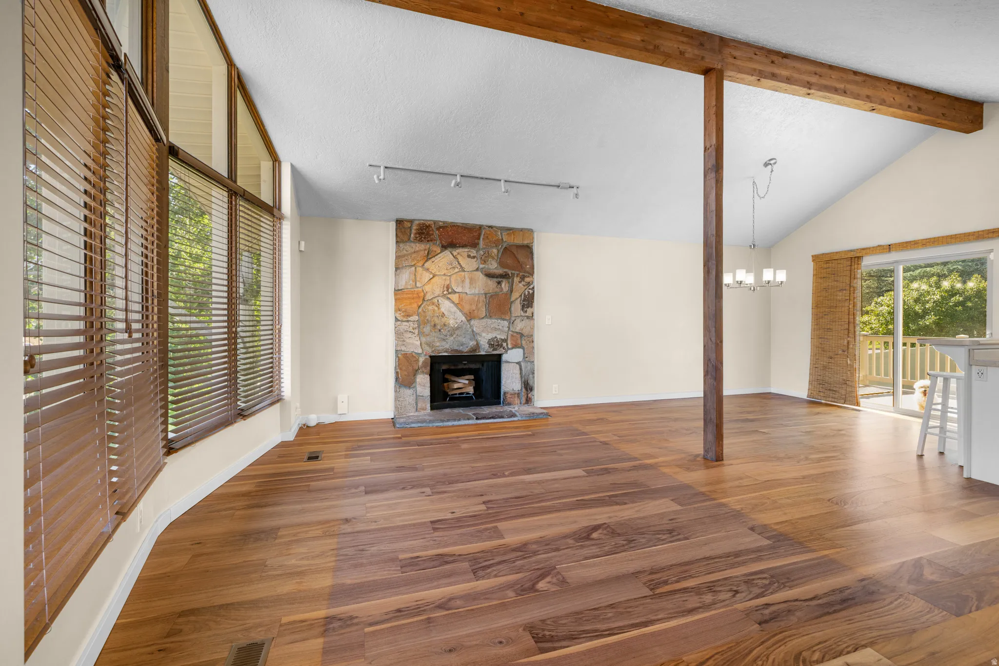 Spacious living area featuring vaulted ceilings, exposed wood beams, a stone-clad fireplace, extensive wood-finish flooring, and large windows with integrated blinds