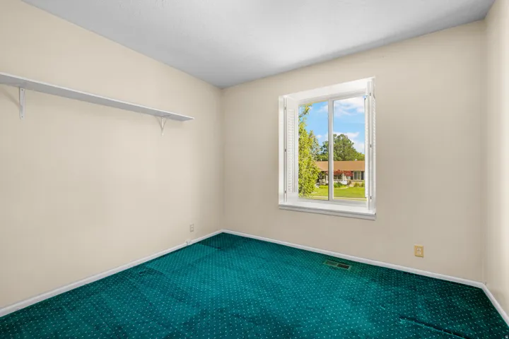 Room featuring a white-framed window with louvered shutters, light beige walls, and teal patterned carpeting