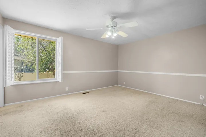 Spacious room featuring neutral carpeting, light beige walls, white chair rail molding, a large window with white plantation shutters, and a ceiling fan with integrated lighting