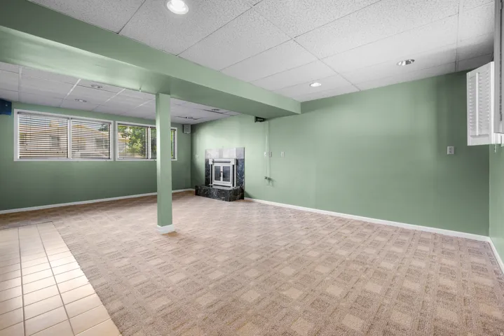 Spacious room featuring a corner fireplace with a dark stone surround, multiple windows with blinds, recessed lighting, and a combination of patterned carpet and tile flooring