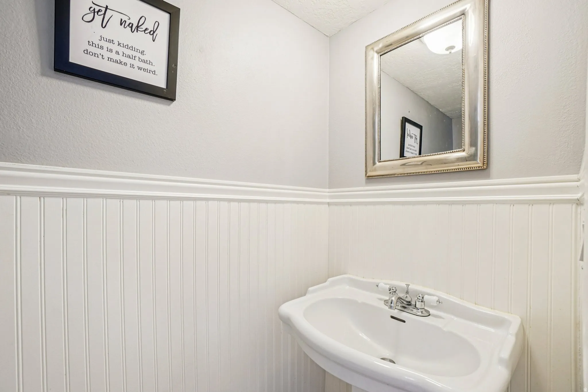 Pedestal sink with chrome faucet, complemented by white beadboard paneling and a decorative wall mirror
